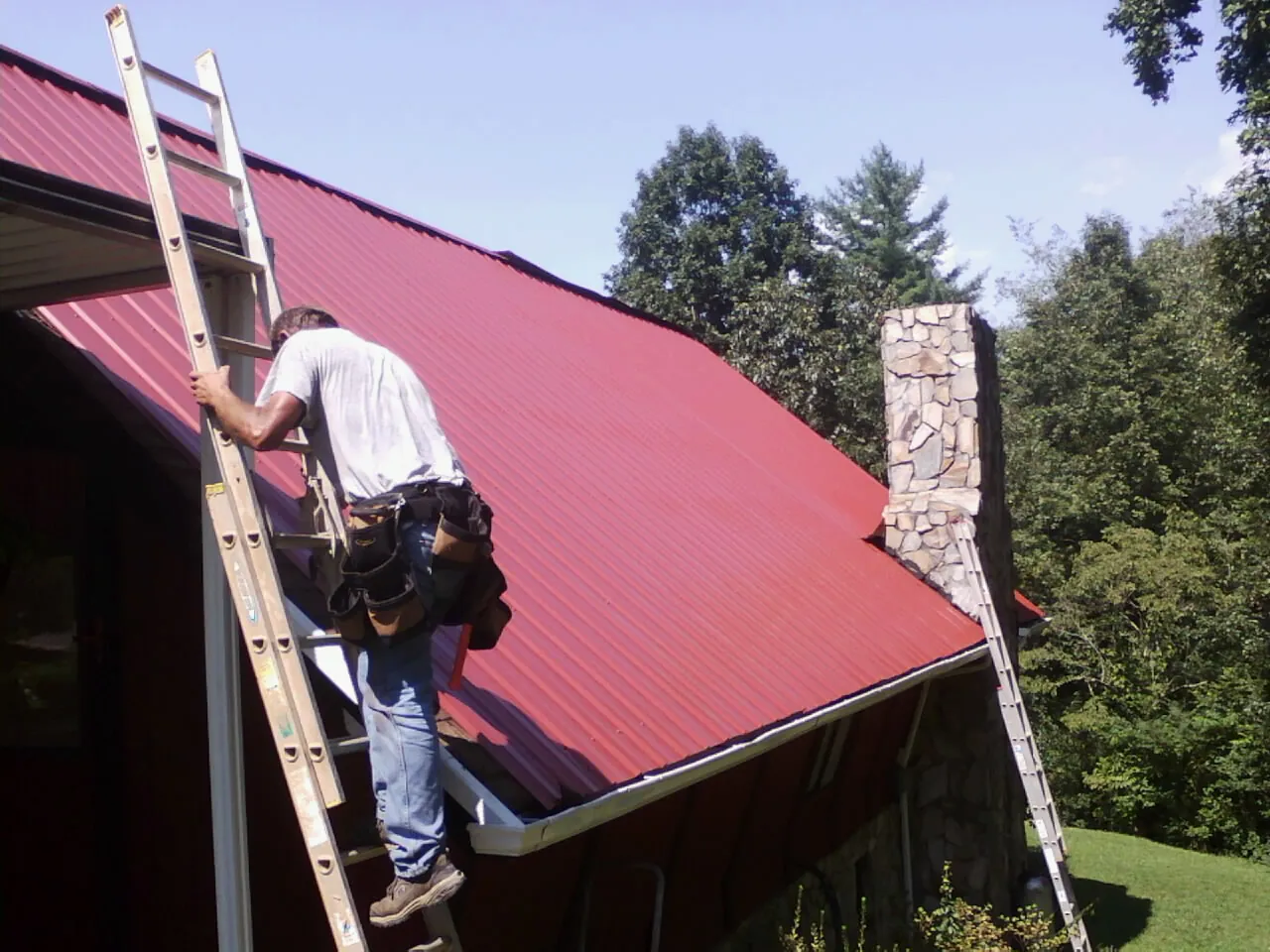 Roofer on a ladder beside a red metal roof and stone chimney — Soffit & Fascia Repair by Roofing Brothers in Staley